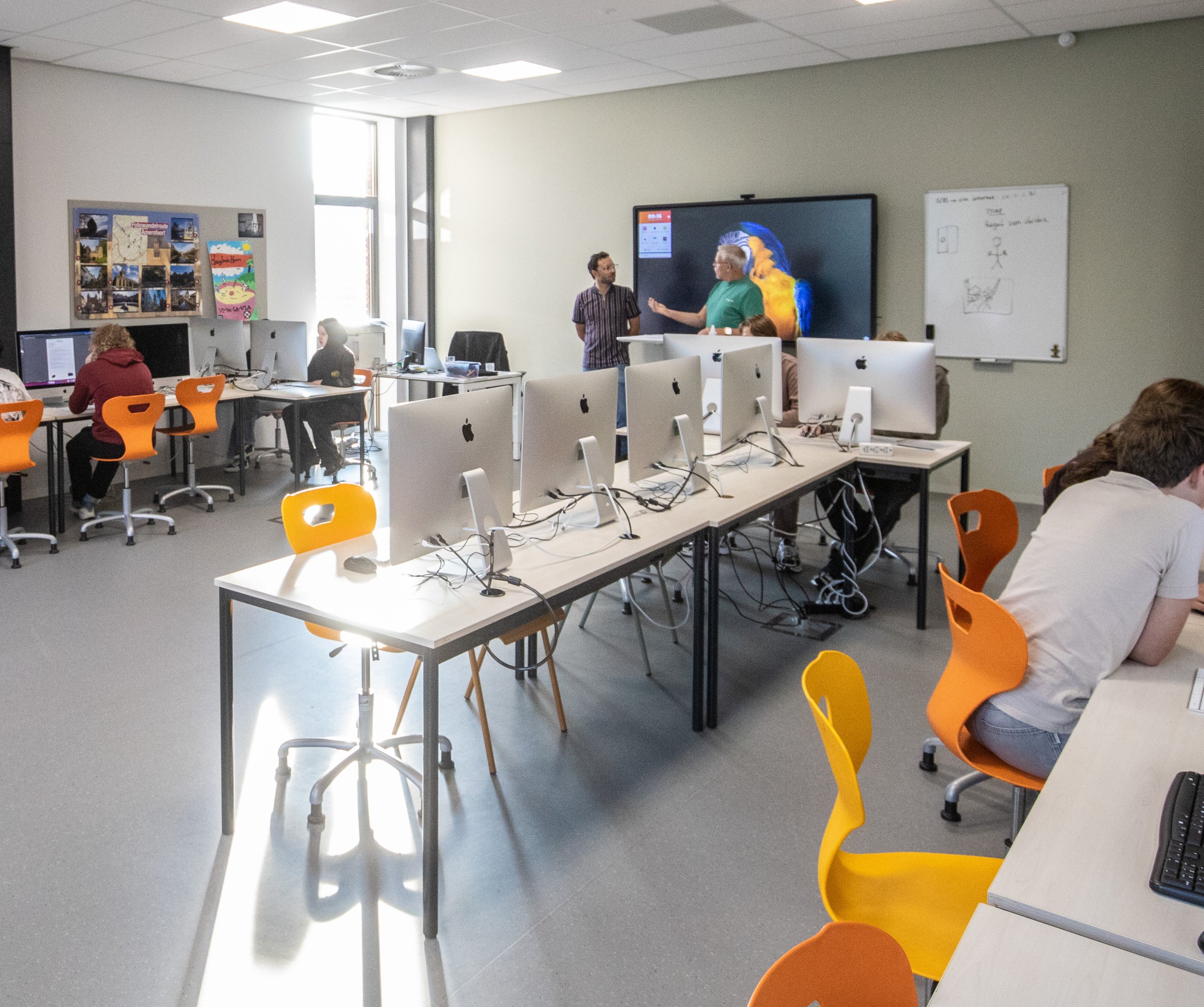 Leerlingen werken achter computers in het computerlokaal, twee docenten bij het digibord