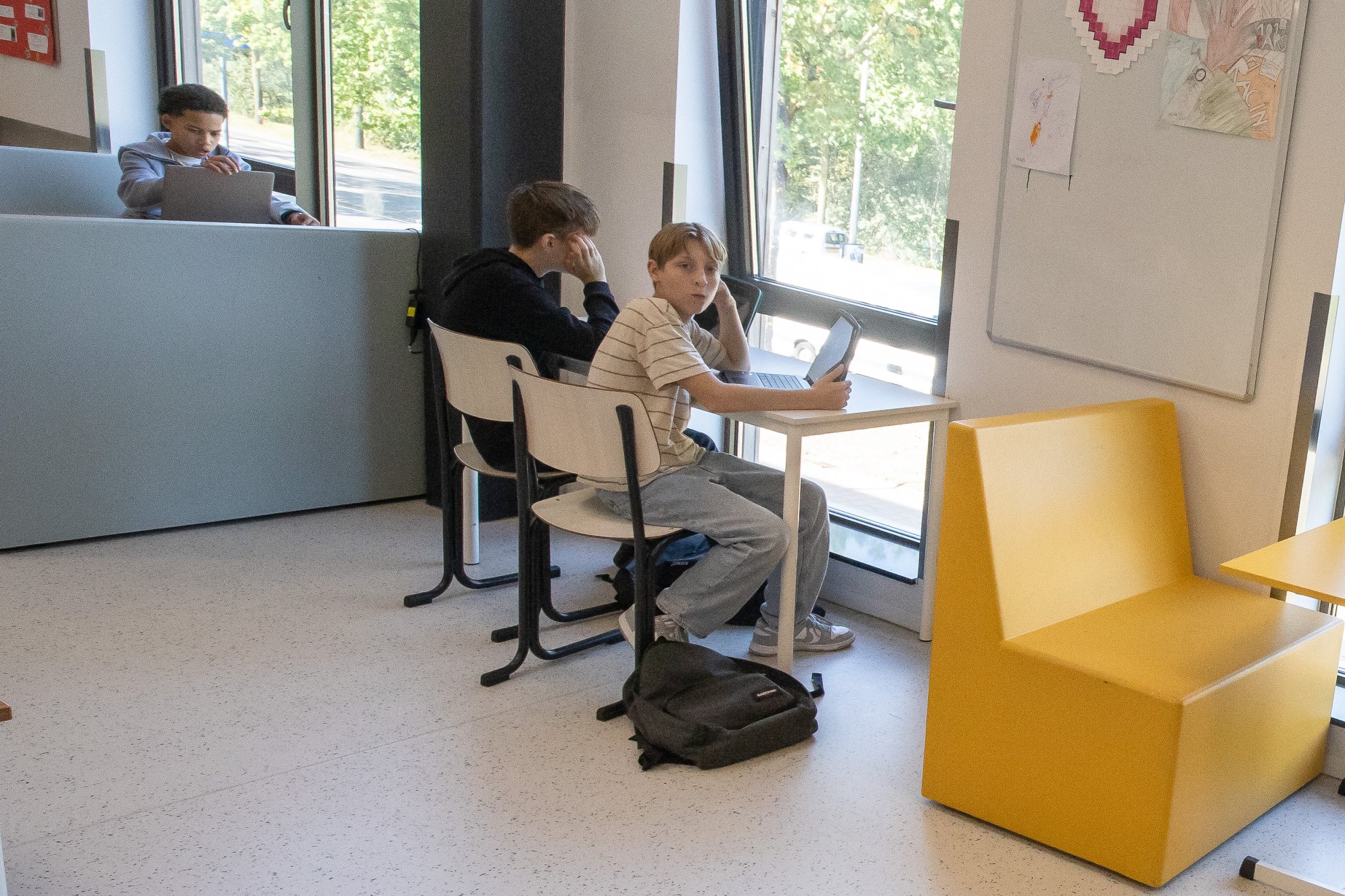 Twee leerlingen werken aan een tafel bij het raam, een derde werkt op een laptop