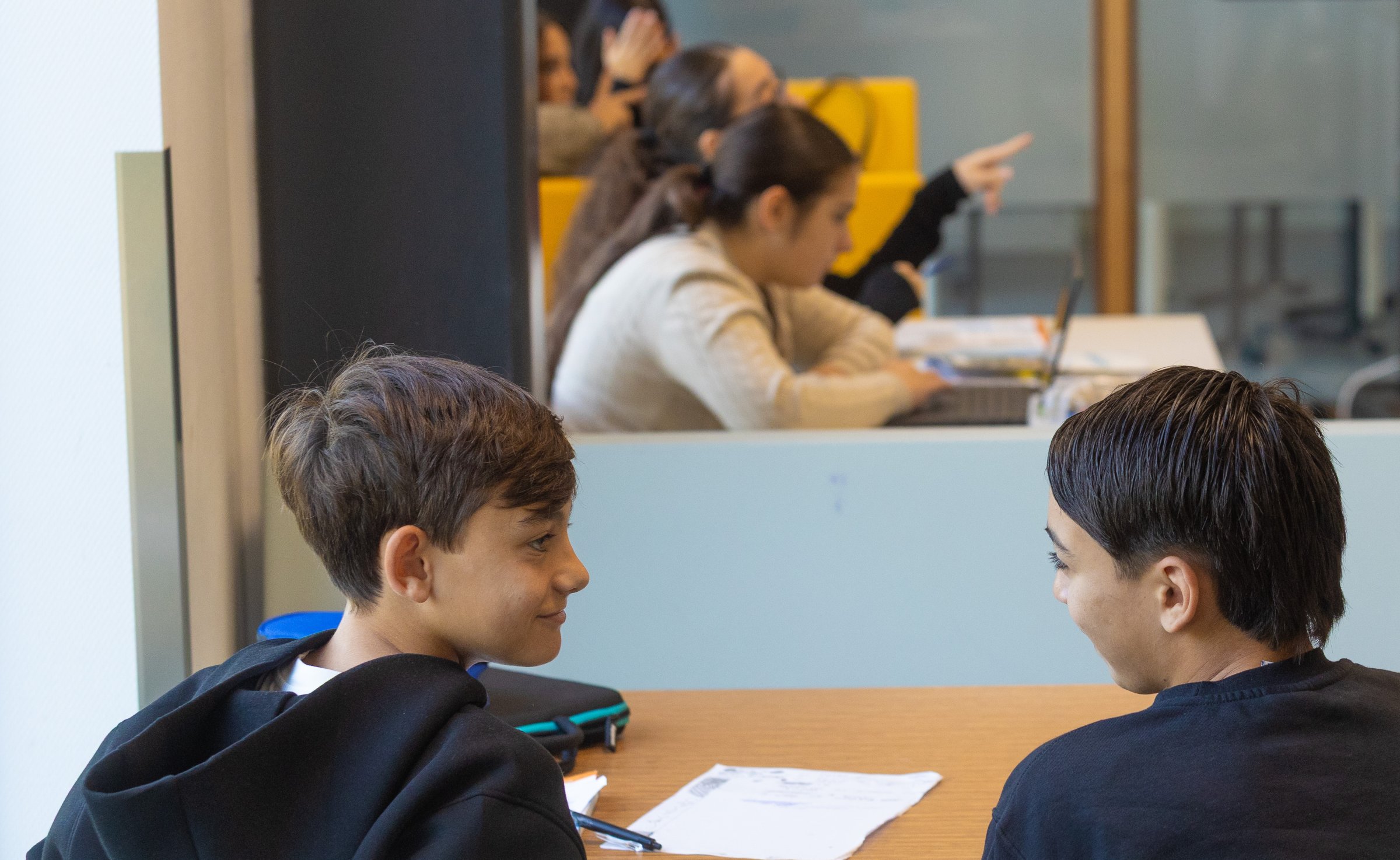 Twee leerlingen kijken elkaar lachend aan aan een tafel met werkbladen